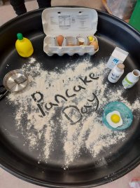 Black tray with wooden play food eggs, milk and lemon. Pancake day is written in spilt flour