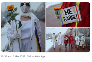 Six women in Belarus walk together, each holding a sunflower. They all wear scarves and glasses. Two women wear red, four wear white. One woman holds a sign that says we don't want this war in Belarusian. 