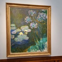 A painting in a gold frame of waterlilies and agapanthus, very brushy and loose in style. The colours are mostly murky but with some glimpses of brighter colour here and there.