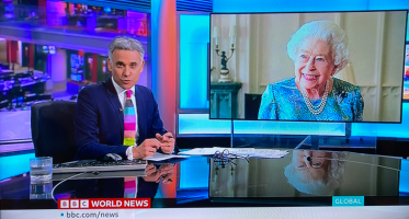 Queen grins maniacallyon screen, eyes toward news man in foreground.