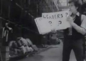 Bob Dylan in a fuzzy still from the Subterranean Homesick Blues short. He is holding a placard which says “leaders ???”