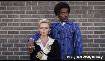 Ruby Sunday and the Doctor. Ruby is blonde with a cream blouse and black jacket. The Doctor has longer Afro hair and a bright blue pin striped suit with wide lapels. 