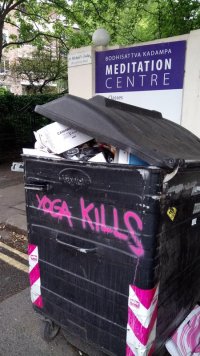 An industrial sized wheelybin overflowing with rubbish. In the background is a sign for the Buddhist meditation centre in the building behind. Someone has spraypainted YOGA KILLS on the side of the bin