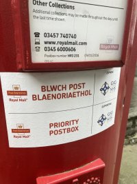 A red pillar box has a bilingual notice in Welsh and English. It’s a priority post box. The paint job looks recent. 