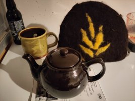 A medium sized dark brown teapot made in the classic round teapot shape, next to a tea cosy made of thick felted wool in the same colour with a contrasting leaf-shaped design