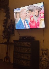 Antiques Roadshow in on the TV. A chest of drawers decorated by Mrs LR is underneath it. There’s a brass side table with a brass Edwardian lamp with a rococo mirror behind it. To the left is a door surrounded by a hop garland. 