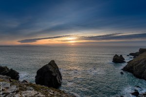 The sun setting over a calm sea with dark rocks in the foreground.