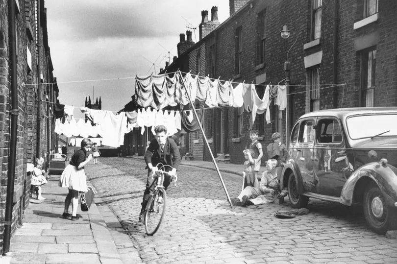 boy riding a bike with various kids playing in a cobbled terraced street in Manchester, which has washing strung across the street.  date unknown