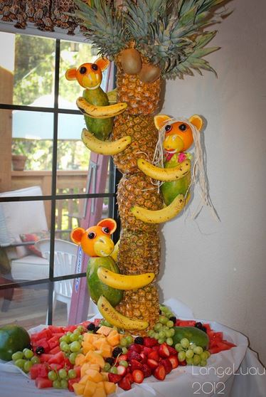 table with four pineapples stacked on top of each other vertically to make a tree trunk, with three monkeys made out of fruit clinging to it.  The base of the table is covered in fruit